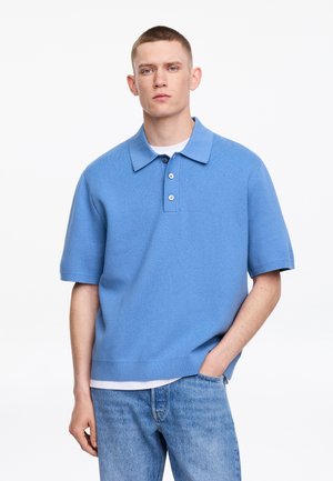 Homme aux cheveux courts portant un polo bleu à manches courtes par-dessus un sous-vêtement blanc et un jean bleu, debout avec une main dans la poche.