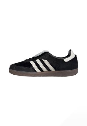 Zwarte lage sneaker met witte strepen, witte hieltab en grijze, gestructureerde zool, zijaanzicht op een witte achtergrond.
