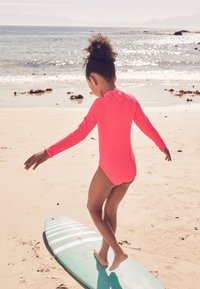 Lichtroze badpak met lange mouwen, gedragen door een kind dat op een turquoise surfplank loopt op een zandstrand, met oceaangolven en zonverlicht water op de achtergrond.