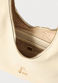 Beige synthetische handtas met ritssluiting, voorzien van een logo in goudkleur en een binnenvoering bedrukt met de merknaam "GUESS".