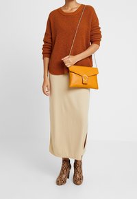 Sweter w kolorze rdzy, beżowa prążkowana spódnica z bocznymi rozcięciami oraz brązowe botki na obcasie z motywem węża. Nosi żółtą torebkę na ramię z okrągłym akcentem.