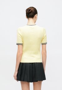 Κίτρινο πλεκτό πουλόβερ με υφή σ χεδίου και κοντά μανίκια, με μοβ λεπτομέρειες στο γιακά και στις μανσέτες, σε συνδυασμό με μαύρη πλισέ φούστα.