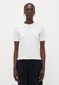 White cotton t-shirt with short sleeves, round neckline, and three decorative gold buttons on the left shoulder. Smooth texture, fitted design.