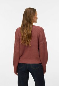 Rokdarbu džemperis netīrā rozā krāsā ar ribveida faktūru un pārāk lielām piedurknēm, kombinēts ar tumši zilām džinsa biksēm, skatīts no aizmugures.