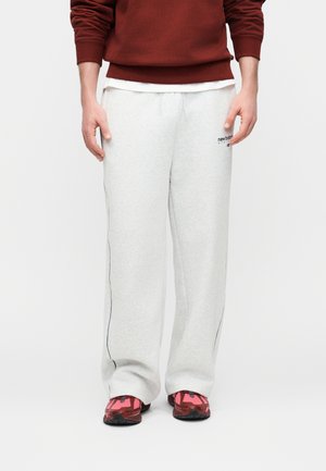 Mees, kes kannab heledat halli laiade säärtega dressipükse küljeribadega, roostevärvi pusa ja punaseid spordijalatsid, seisab ühtlase tausta ees.