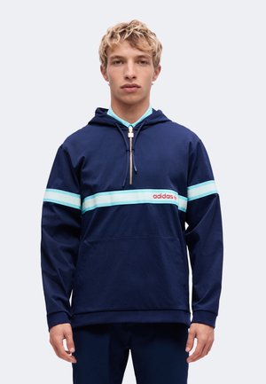 Ung mand iført en mørkeblå Adidas hættetrøje med lyseblå striber og en lynlås foran, stående mod en ensfarvet lys baggrund.