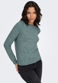 Žalias megztinis suapvalintu kaklu ir ilgomis rankovėmis, turintis tekstūruotą paviršių. Derinamas su juodomis džinsinėmis kelnėmis.