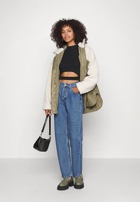 Mulher com cabelo encaracolado, vestindo um top cropped preto, jaqueta verde com mangas brancas, jeans azuis, botas verdes e segurando uma bolsa preta.