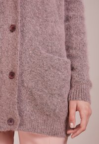 Stickad kofta i mjuk rosa, med runda bruna knappar, ribbade ärmslut och två framfickor med en mysig, strukturerad finish.