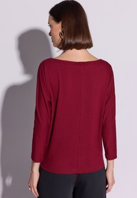 Bordowa bluzka z długim rękawem o szerokim dekolcie, z prostym krojem i przeszyciem z tyłu, wykonana z miękkiego, lekkiego materiału.