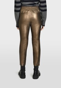 Metalické bronzové slim-fit kalhoty s elastickým pasem, zadními kapsami a hladkým povrchem, v kombinaci s černými robustními botami.