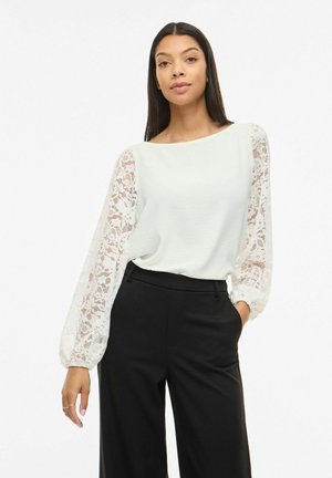 Kobieta w białej bluzce z przezroczystymi koronkowymi długimi rękawami i czarnych spodniach z wysokim stanem, stojąca z jedną ręką w kieszeni na białym tle.