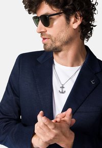 Granatowa marynarka z fakturowanej tkaniny, biała koszula, srebrny naszyjnik w kształcie kotwicy i czarne okulary przeciwsłoneczne. Ręce złożone, skupione na biżuterii.