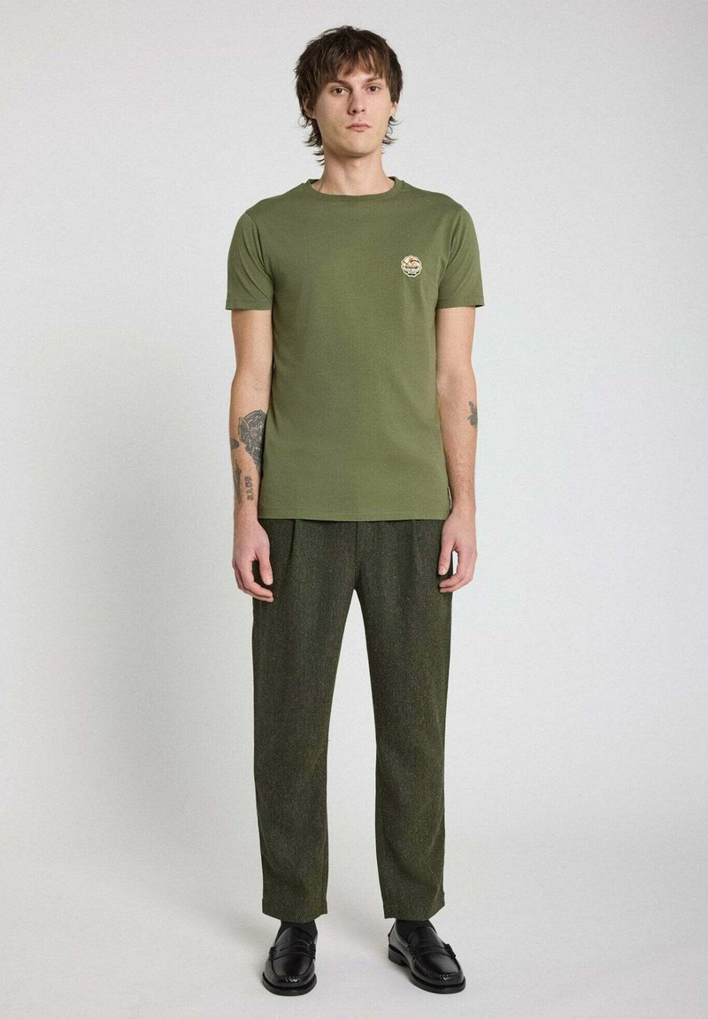 T-shirt vert olive à col rond avec un petit écusson sur la poitrine, associé à un pantalon texturé vert foncé et des chaussures noires.