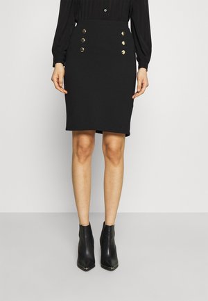 Jupe crayon noire en tissu extensible, dotée d'une taille haute et de quatre boutons dorés à l'avant. Associée à des bottines noires.