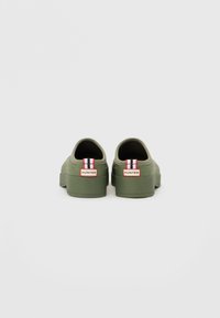 Zöld gumí slip-on cipők lekerekített szélekkel, lapos talppal és piros, fehér és fekete csíkos díszítéssel a saroknál. "HUNTER" logó részlet.