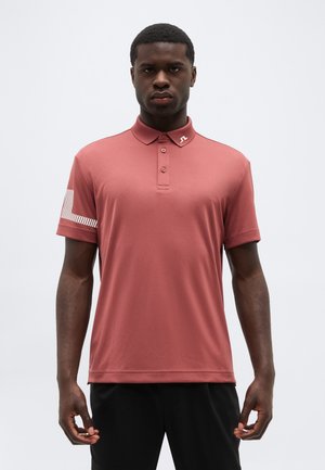 Man wearing a muted red polo shirt with three buttons and a white geometric stripe on the left sleeve, standing against a plain background.