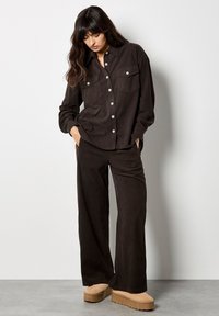 Bruine ribfluwelen shirt met twee voorzakken en zilveren knopen, gecombineerd met bijpassende wijde pijpenbroeken en tan platform schoenen.