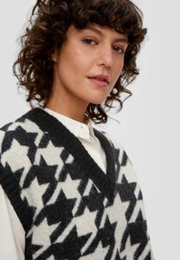 Gilet en tricot noir et blanc avec un col en V, présentant un motif étoile. La texture est douce, avec des bords côtes et une coupe ample.