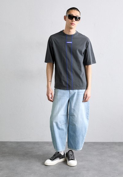 Gray cotton t-shirt with short sleeves, featuring blue stitching down the center. Worn with loose light blue jeans and black sneakers.