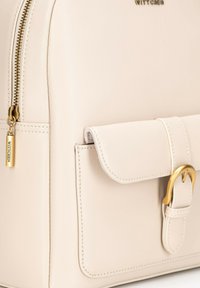 Beżowy skórzany plecak z zamkiem błyskawicznym, przednią klapą z detalem w postaci klamry oraz subtelnymi akcentami ściegów. Widoczna złota metka.