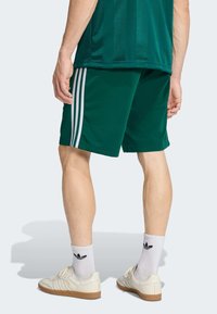 Zelené športové šortky s tromi bielymi pruhmi na každej strane, doplnené bielymi teniskami a pruhovanými ponožkami, zobrazené zozadu.