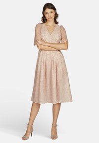 Spitzenkleid in hellrosa mit V-Ausschnitt, kurzen Puffärmeln und taillierter Taille. Ausgestellter Rock mit gewelltem Saum und aufwendigem Muster.