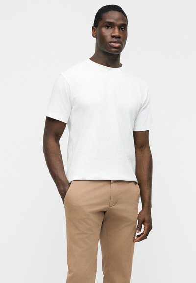 Jeune homme portant un t-shirt blanc à manches courtes uni et un pantalon marron clair, debout devant un fond blanc uni.
