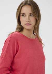 Pull corail présentant un tissu doux et texturé, un col rond et des épaules tombantes. Les manches sont ornées de détails côtelés pour plus de style.