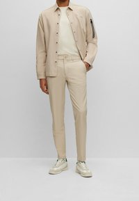 Beige jacka med en dragkedjeficka och knappar, tillsammans med ljusfärgade slim-fit byxor och beige sneakers med gröna detaljer.