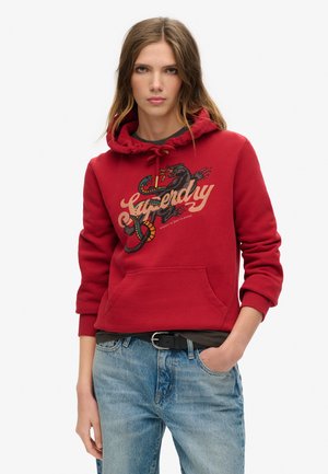 Sudadera roja con capucha, con un gráfico de una serpiente y el texto "Superdry". Presenta un bolsillo tipo canguro y puños acanalados. Combinada con jeans azules.