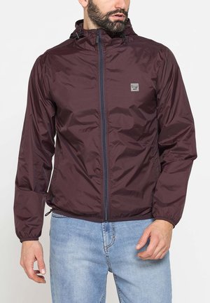 Uomo che indossa una giacca con cappuccio bordeaux scuro con cerniera e polsini elasticizzati e jeans azzurri su sfondo neutro.