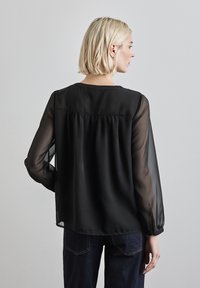 Svart blus med långa genomskinliga ärmar, rynkad detalj på baksidan och rund halsringning, tillsammans med mörka denimjeans. Slät textur.