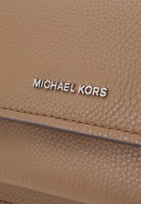 Borsa a tracolla in pelle color cuoio con superficie texturizzata. Presenta un logo argentato "MICHAEL KORS" e cuciture visibili lungo i bordi.