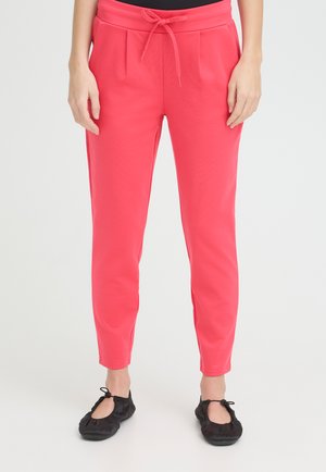 Femme portant un pantalon fuselé rose vif avec une taille à cordon et des ballerines noires sur un fond uni.