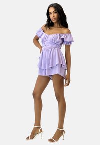 Vestido lila de hombros descubiertos con mangas de volantes, diseño en capas y cintura ajustada, combinado con tacones plateados de tiras.