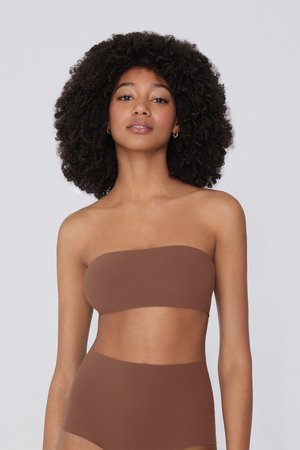 Frau mit lockigem Haar, die ein schulterfreies braunes Bandeau-Oberteil und hochtaillierte braune Unterteile trägt, steht vor einem schlichten Hintergrund.