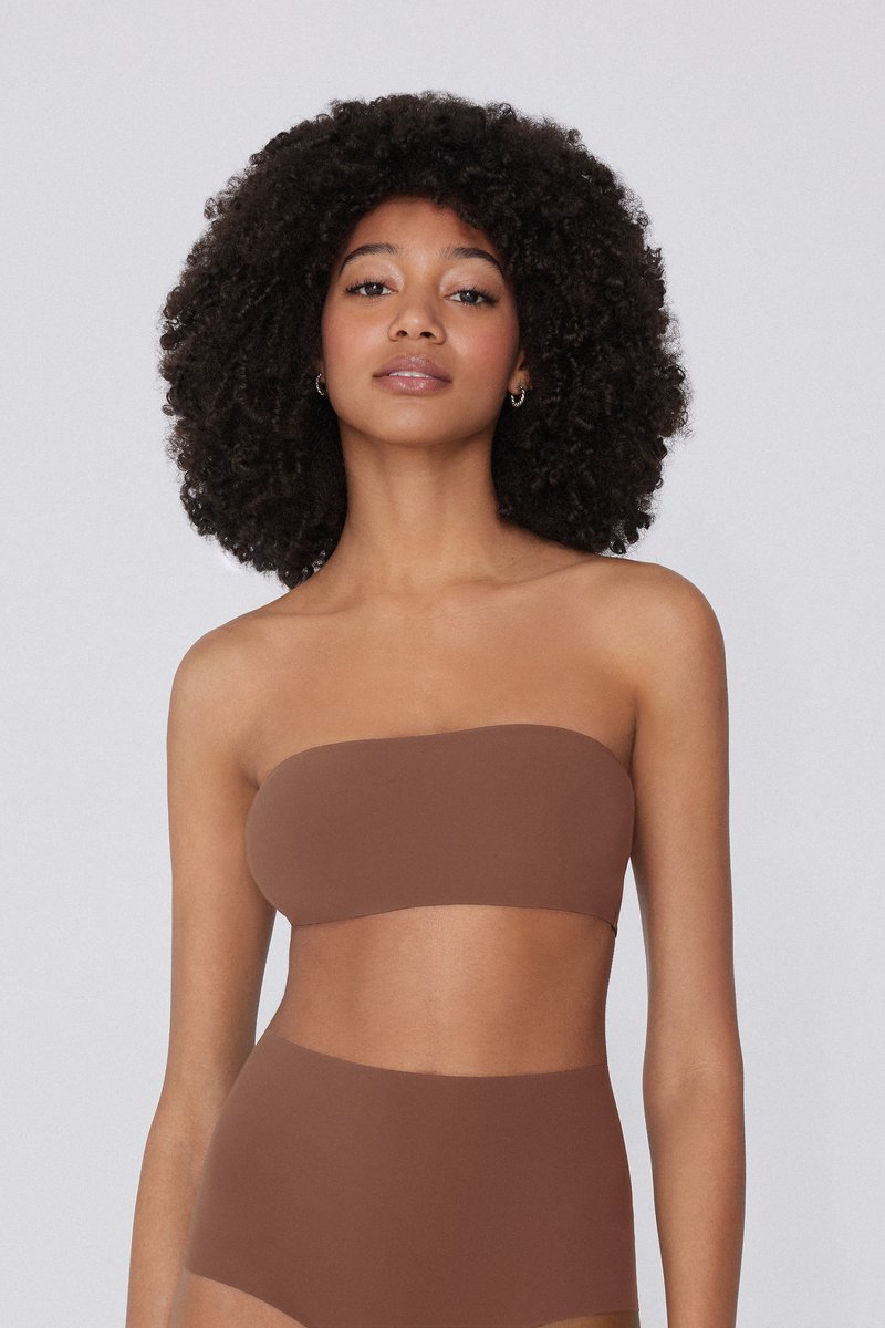 Mujer con cabello rizado, vestida con un top bandeau marrón sin tirantes y pantalones marrones de talle alto, de pie contra un fondo liso.