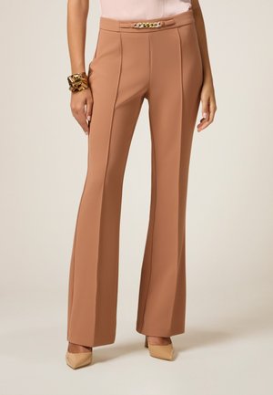 Pantalons évasés beiges avec une texture lisse, dotés d'un accent de chaîne en or à la taille, associés à des talons nude.