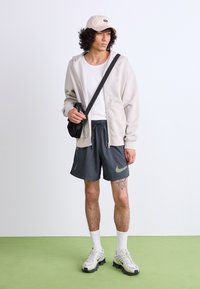 Lys beige zip-up hoodie, hvid tank top, mørkegrå shorts med grønt Nike-logo, hvide sokker og grå sneakers med sorte detaljer.