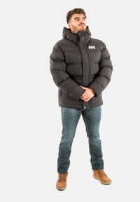 Giacca a vento nera con colletto alto e cappuccio. Presenta una chiusura con zip, cuciture orizzontali e un logo sul petto. Indossata con jeans blu.