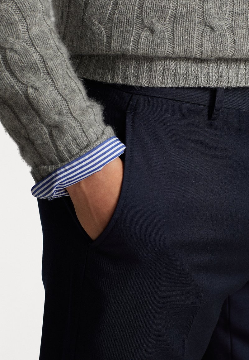 Un pull en maille côtelée gris accompagné d'un pantalon bleu marine foncé. Le poignet de la chemise présente des rayures bleues et blanches, visibles sous l'ourlet du pull.