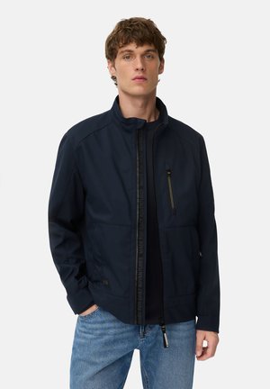 Young man with short brown hair wearing a navy zip-up jacket with chest pocket and light blue jeans against a plain light background.