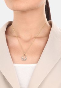 Collana placcata in oro con pendente a goccia decorato con piccoli cristalli trasparenti, indossata sopra un blazer beige chiaro.