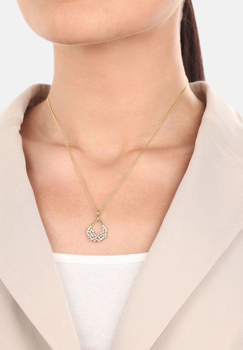 Collana placcata in oro con pendente a goccia decorato con piccoli cristalli trasparenti, indossata sopra un blazer beige chiaro.