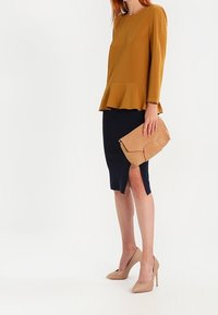 Top peplum senape, gonna a tubino blu navy con spacco laterale, tacchi alti beige e una pochette color cammello. Tessuto liscio e design strutturato.