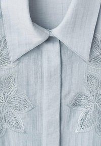 Camisa azul clara con un cuello estructurado, tejido texturizado y bordado de encaje floral detallado en la parte delantera.
