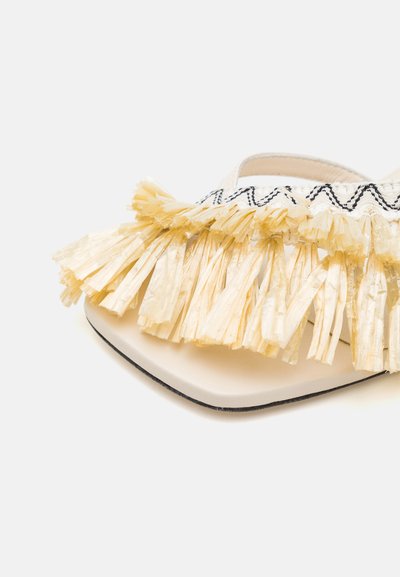 White sandal featuring a large cream fringe accent at the front, with black zigzag stitching above. Square toe design and flat sole.