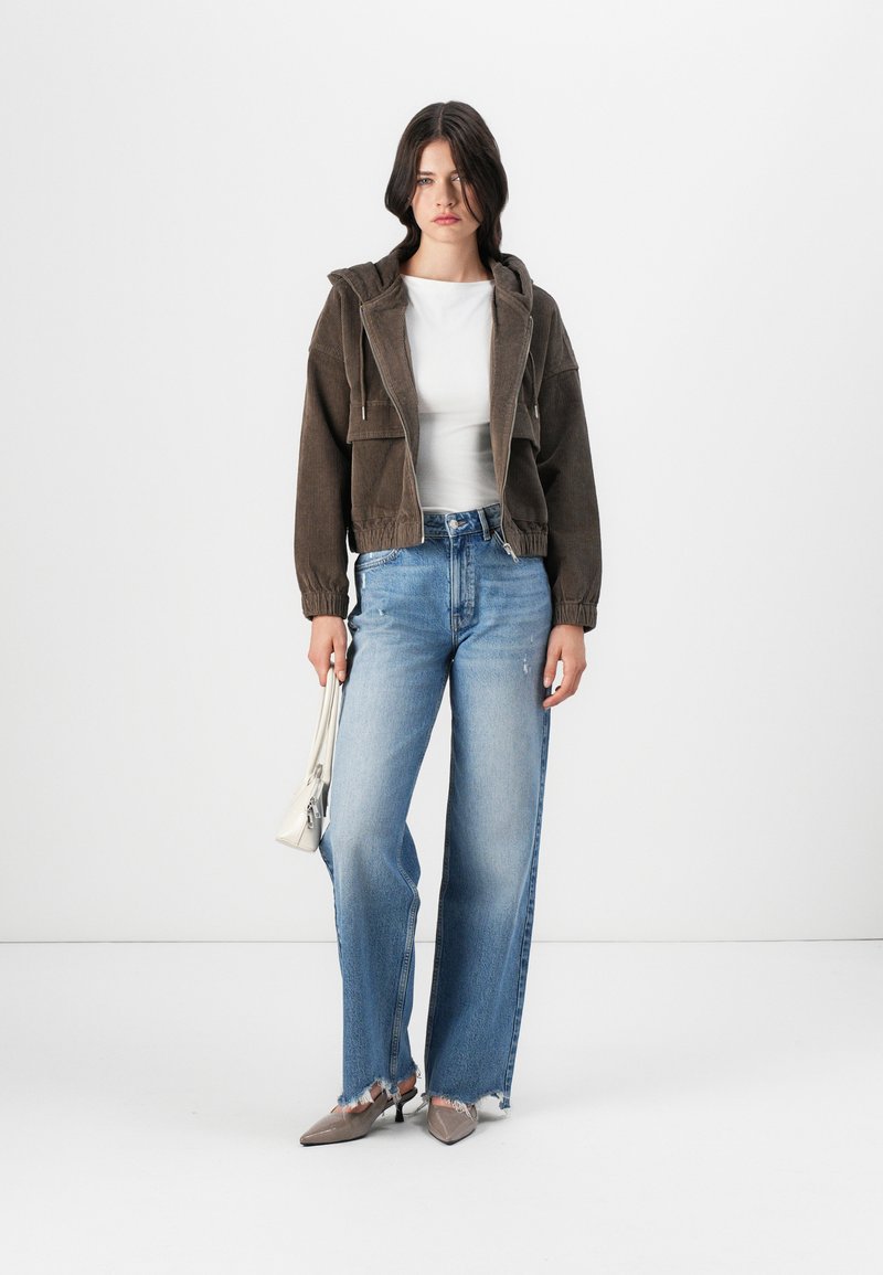 Veste cropped en velours côtelé marron, chemise blanche ajustée, jeans large bleu clair avec ourlets effilochés, et ballerines pointues grises. Tenant un petit sac blanc.