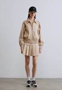 Μπεζ μπουφάν bomber με ελαστικά μανίκια, συνδυασμένο με μια πλισέ φούστα. Η εμφάνιση περιλαμβάνει μαύρα αθλητικά παπούτσια και μαύρο καπέλο, σε ουδέτερο φόντο.